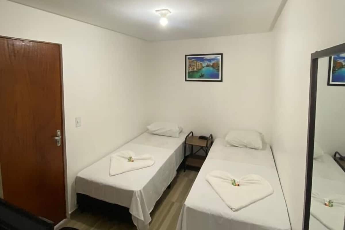 Una habitación pequeña y limpia con dos camas individuales, una puerta de madera, un escritorio con una silla y un cuadro en la pared.