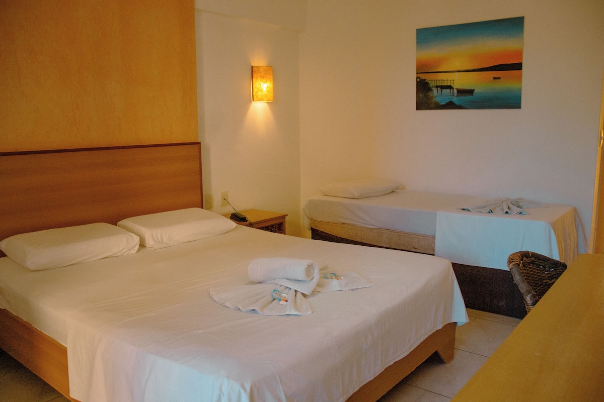 Habitación de hotel con una cama grande, una cama más pequeña, un cuadro de un atardecer y una mesita de noche con una lámpara.