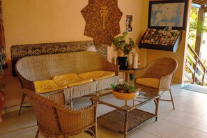 Un sofá de mimbre con cojines amarillos, un sillón de mimbre, una mesa de centro de cristal con una planta, y un mapa y expositor de tarjetas montados en la pared.