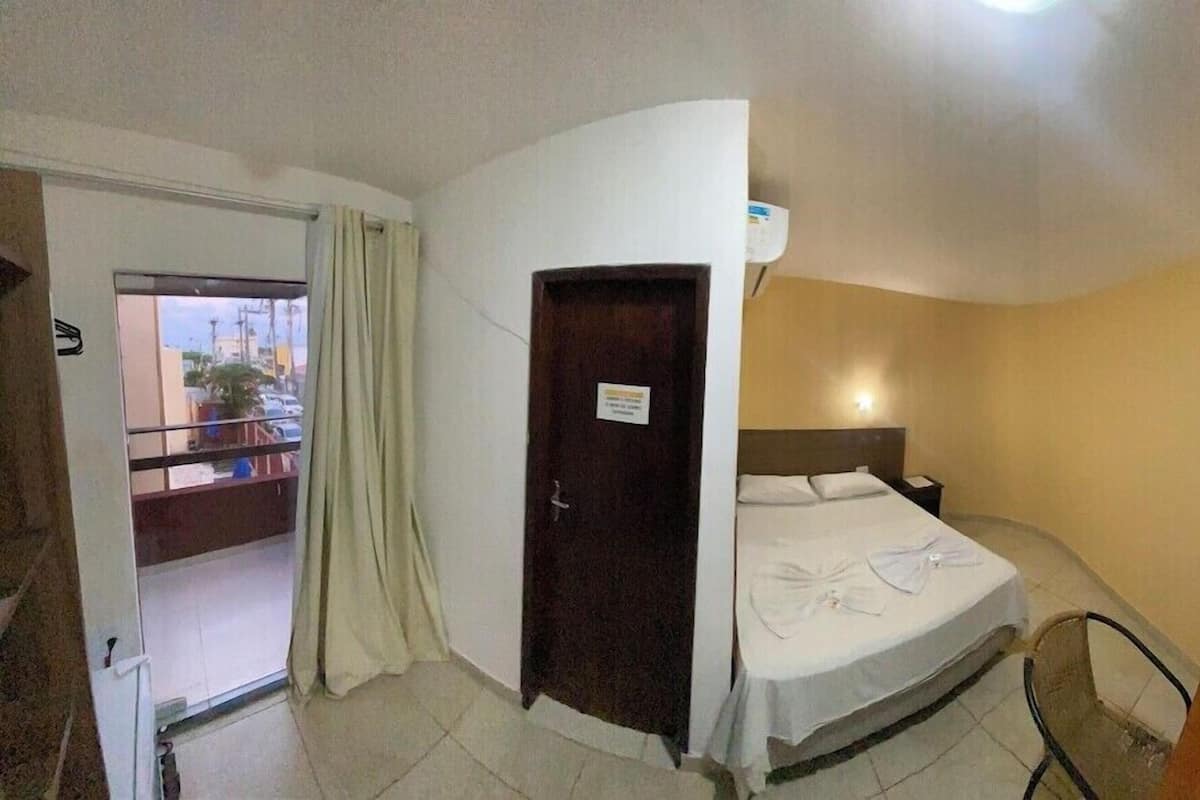 Habitación de hotel con cama, mesita de noche, una silla, una puerta y vistas al exterior.