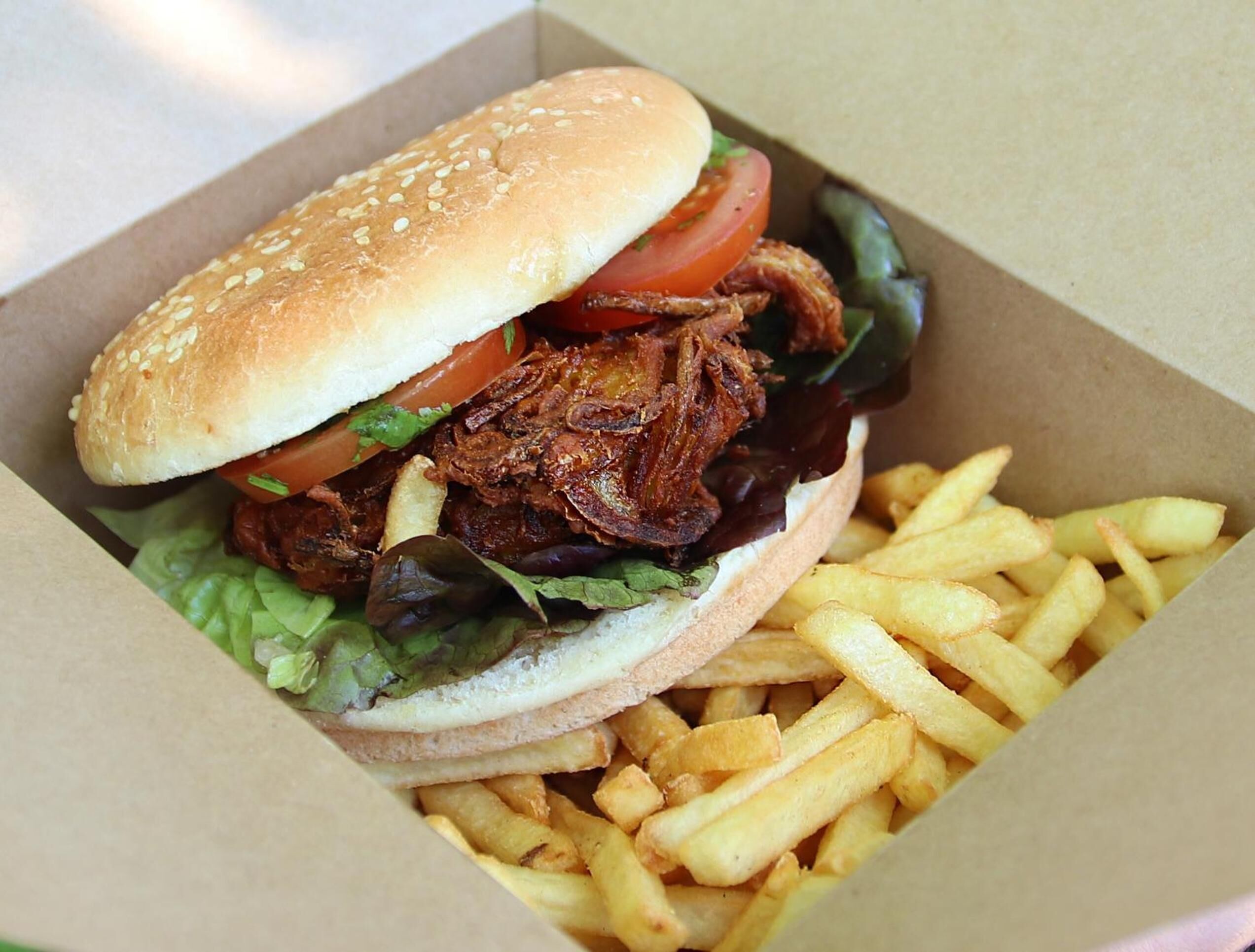 Un sándwich con pan de semillas de sésamo, lechuga, tomate y cerdo desmenuzado, acompañado de papas fritas en un recipiente de cartón.