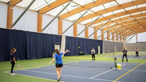 Un court de tennis intérieur avec un filet, des poutres en bois et un fond de scène constitué d’un rideau bleu.