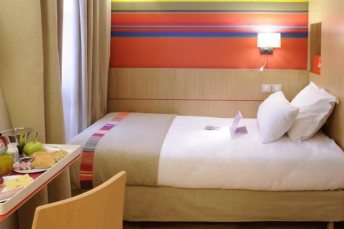 Hotelkamer met een bed, nachtkastje met een dienblad ontbijtartikelen, een stoel en een muur met een kleurrijk streepdessin.