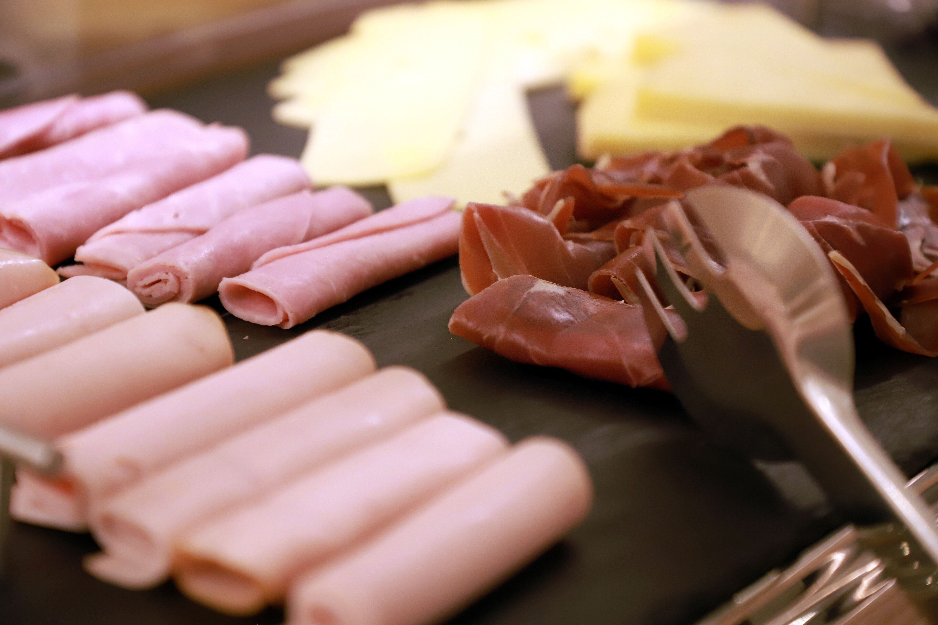 A variety of sliced meats on a board, including ham and possibly prosciutto, with a knife and fork nearby.