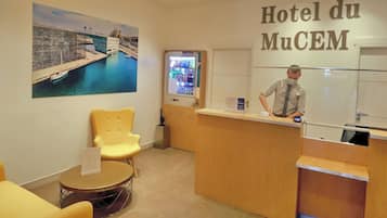 A hotel reception area with a counter, a seating area with yellow chairs, and a large painting of a bridge over water.