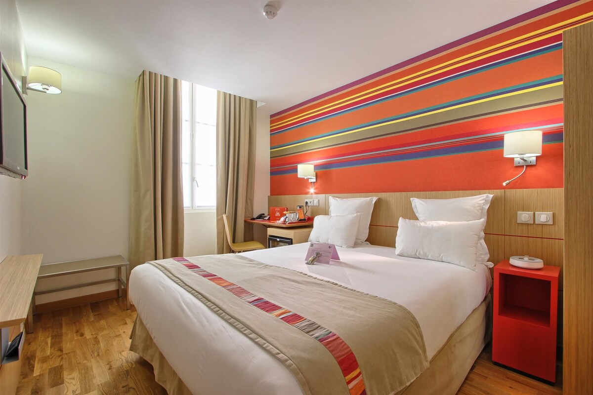 A hotel room with a large bed, a desk, a chair, a nightstand, and a window with curtains.