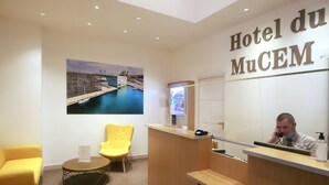 A hotel reception area with a reception desk, a large mirror, a yellow chair, a round table, and a wall-mounted TV screen. - Best Western Hotel du Mucem (Marseille)