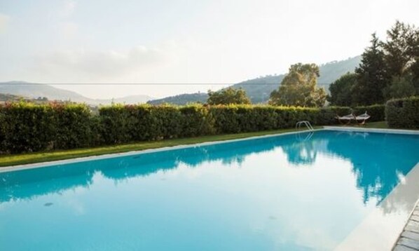 Ein klarer, blauer Swimmingpool, umgeben von ordentlich geschnittenen Hecken und Bäumen, mit Blick auf entfernte Hügel.