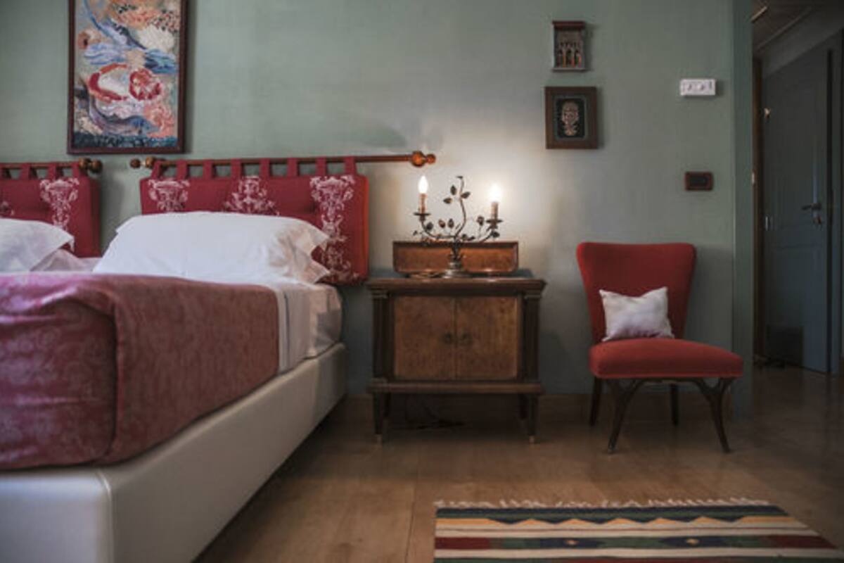 Une chambre à coucher avec un lit, une table de chevet surmontée d’un chandelier, un fauteuil rouge et un tableau accroché au mur.