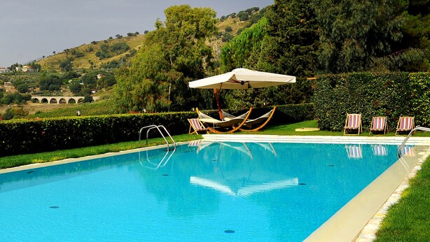 A swimming pool with a hammock and lounge chairs, surrounded by greenery and hills. - Agrirelais Baglio di Pianetto (Santa Cristina Gela)