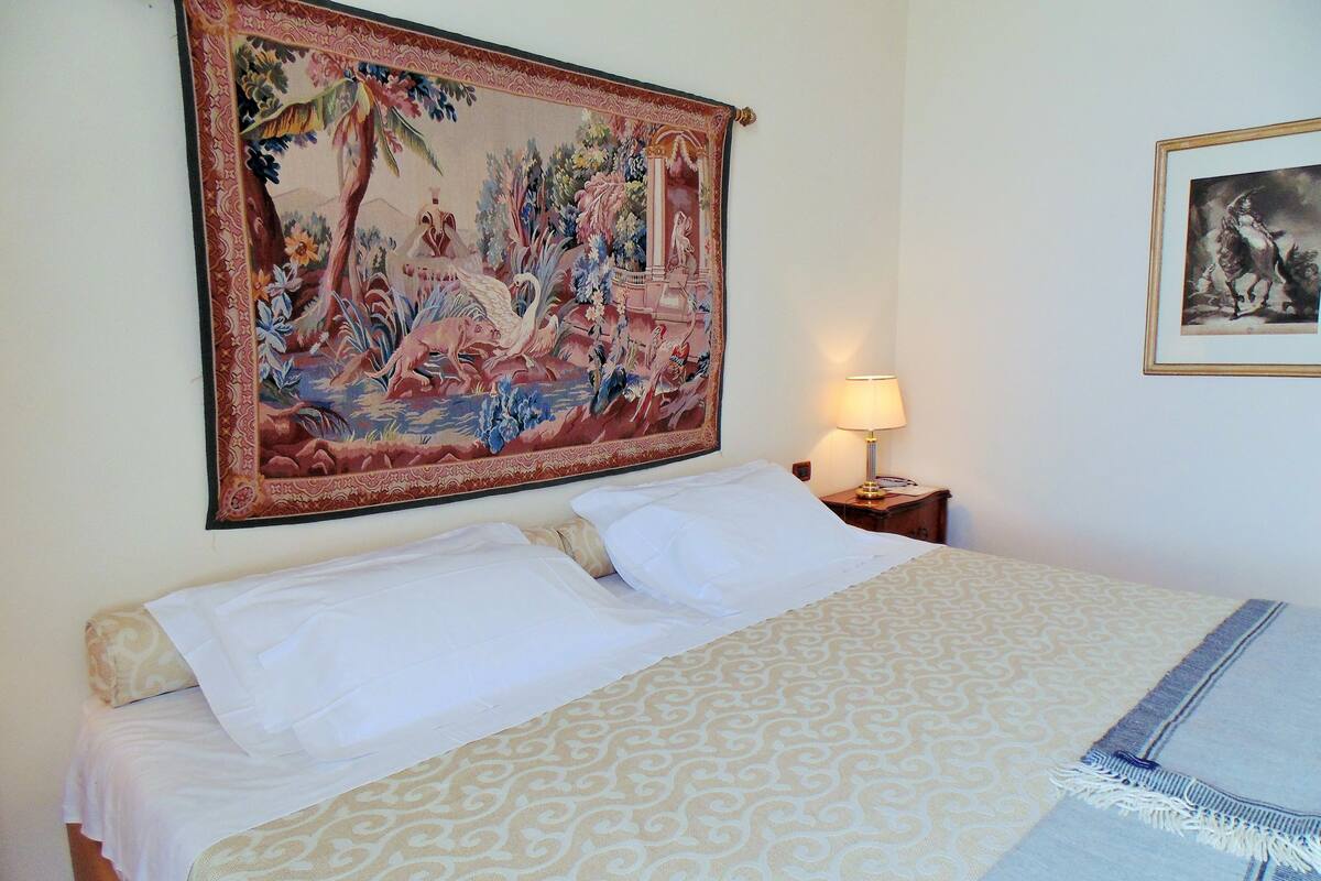 Une chambre d’hôtel avec une grande tapisserie, un lit avec une literie à motifs, une table de chevet avec une lampe et un tableau encadré au mur.