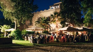 An outdoor gathering with people seated at tables under umbrellas, surrounded by trees and a building with a balcony. - Agrirelais Baglio di Pianetto (Santa Cristina Gela)