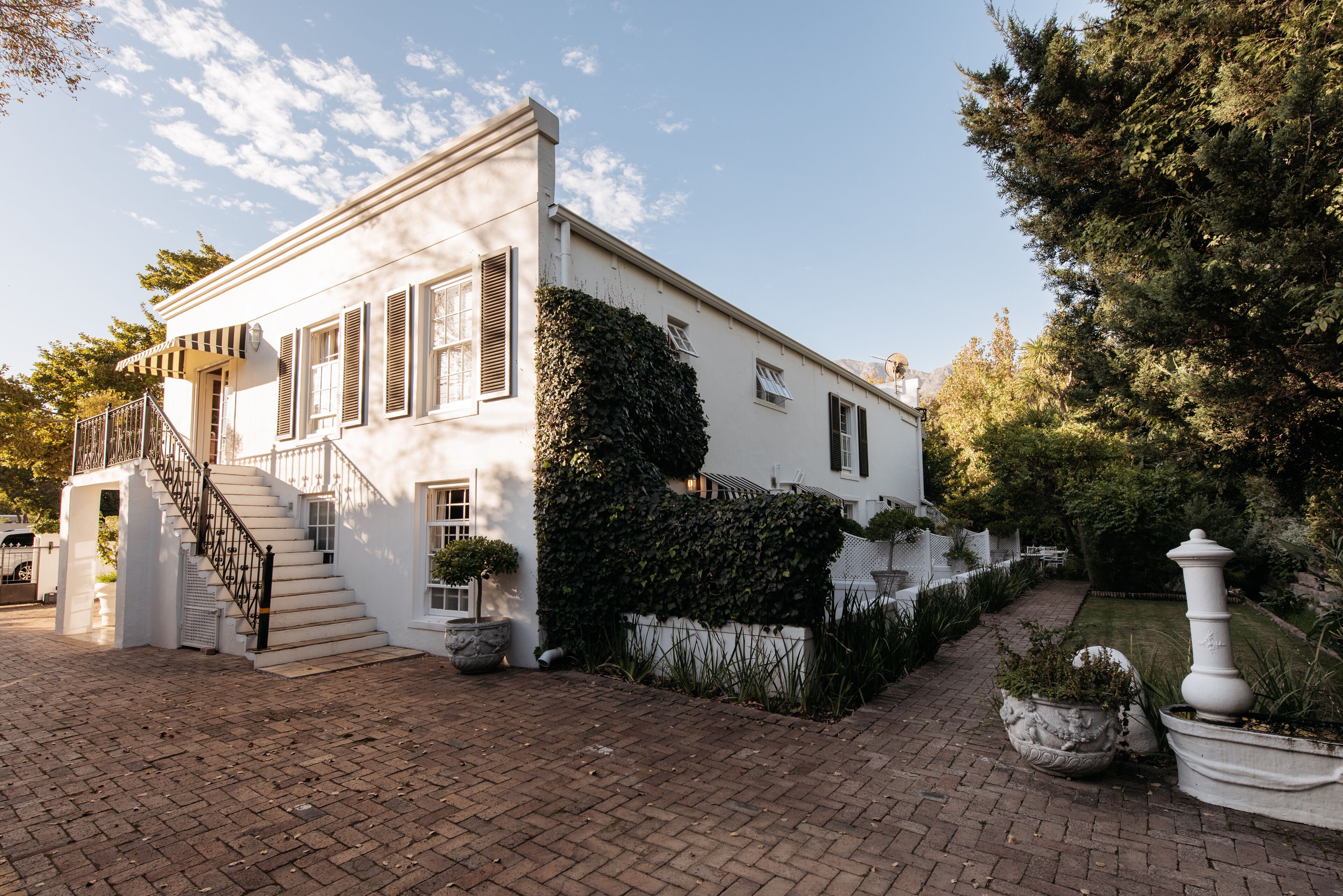 A white building with a brick pathway, a staircase, and a garden area with potted plants.