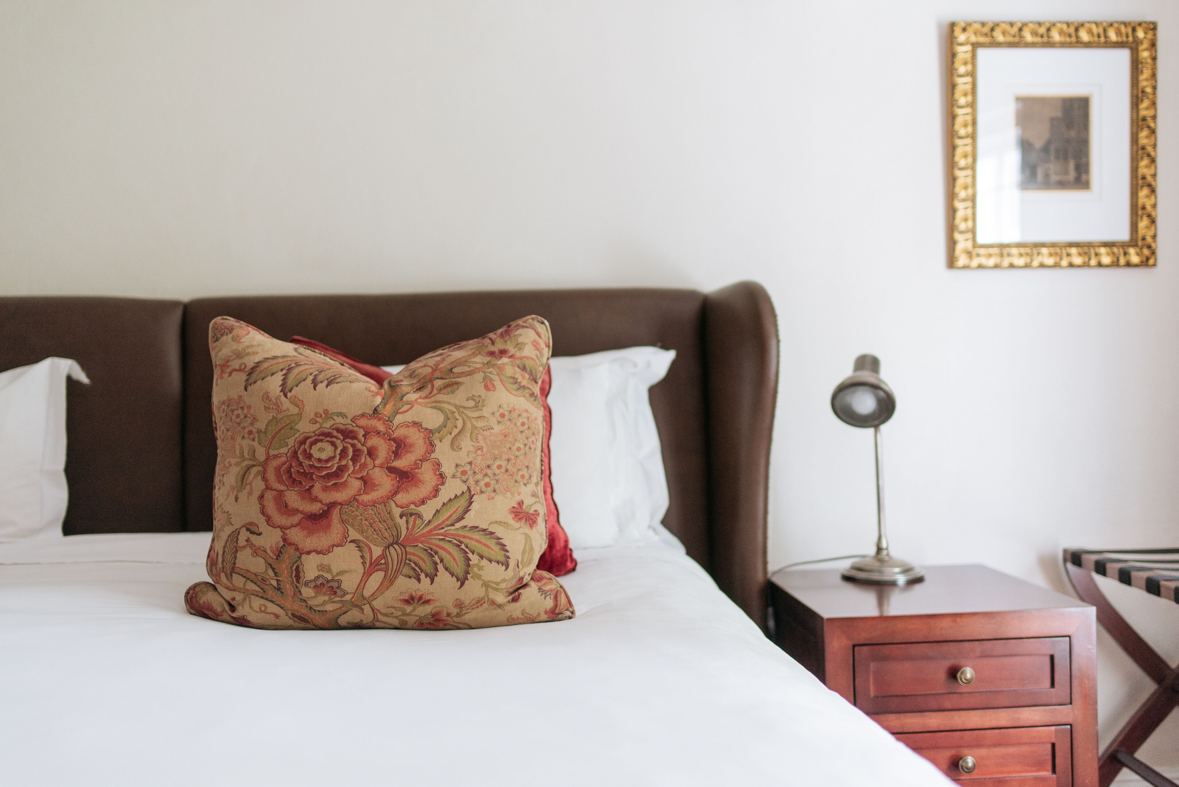A neatly made bed with a floral pillow, a bedside table with a lamp, and a framed picture on the wall.