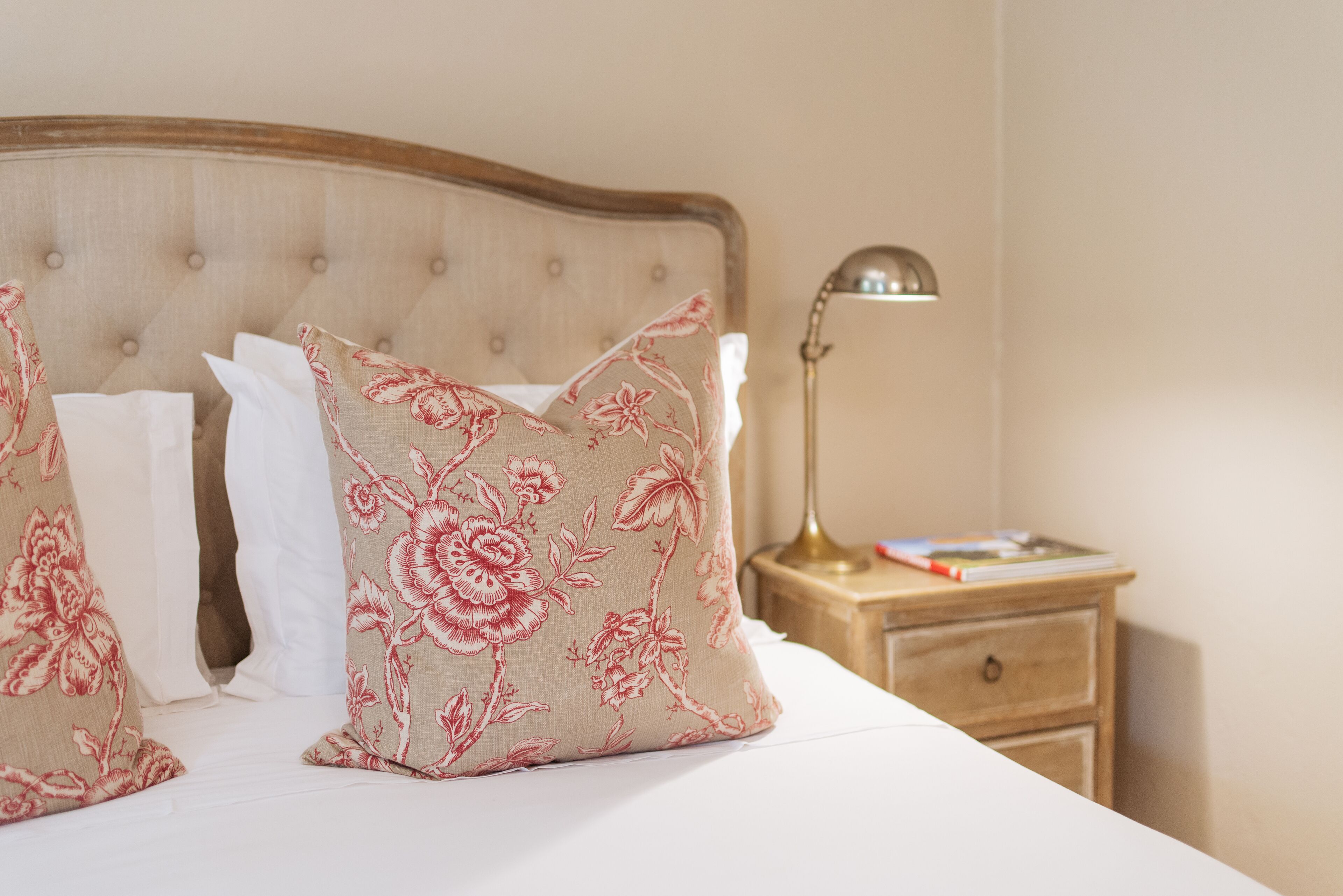 A bed with floral-patterned pillows, a wooden headboard, and a bedside table with a lamp and a book.
