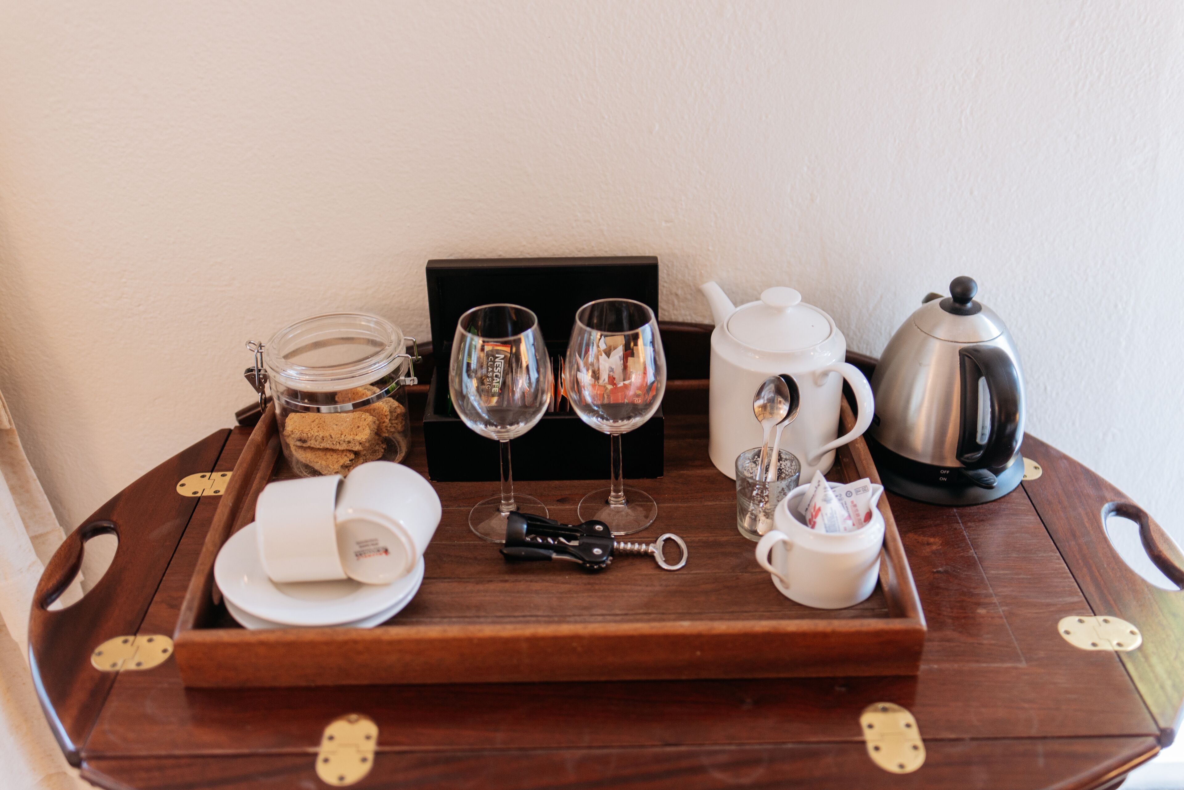 A wooden tray with a jar of cookies, two wine glasses, a teapot, a coffee pot, and a wine opener on a table.