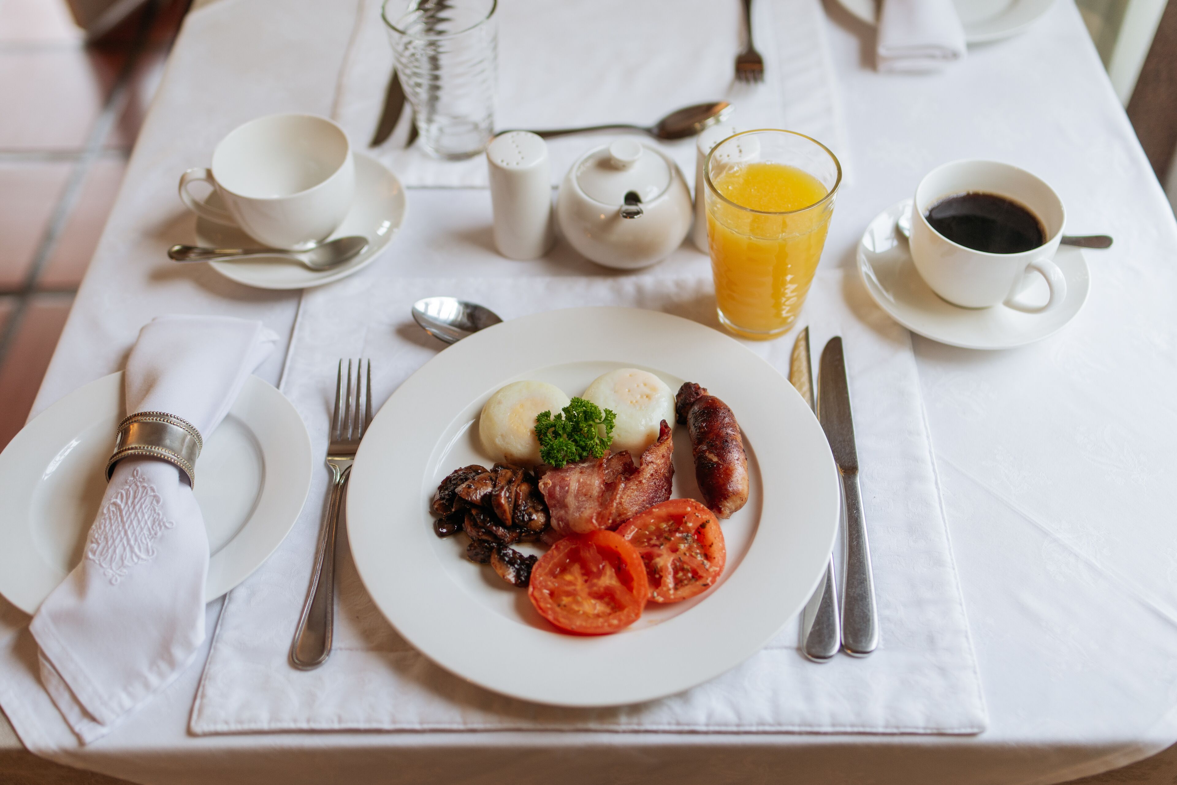 Een ontbijttafel gedekt met borden vol eieren, spek, worstjes, champignons en tomaten, vergezeld van kopjes koffie en sinaasappelsap.