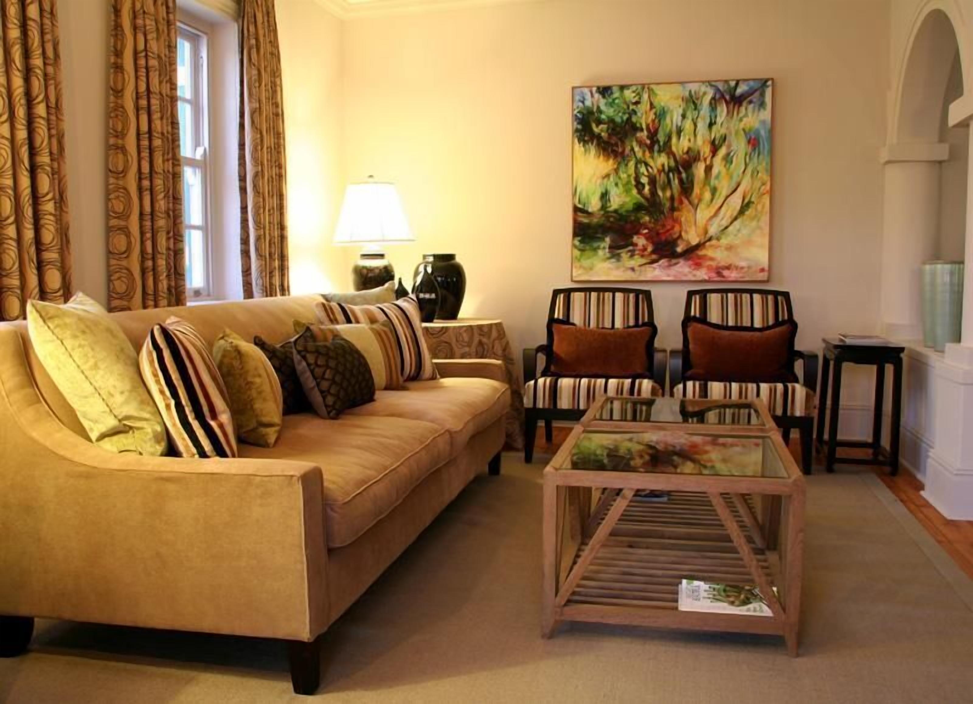 Una sala de estar con un sofá color beige, sillones a rayas, una mesa de centro de vidrio, un cuadro en la pared y una lámpara.