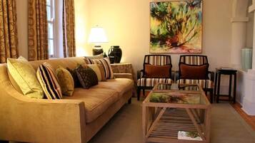 Una sala de estar con un sofá color beige, sillones a rayas, una mesa de centro de vidrio, un cuadro en la pared y una lámpara.