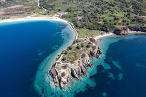 An aerial view of a coastal area with a rocky peninsula, clear blue waters, and a few buildings in the background. - Villa Maria (Sithonia)