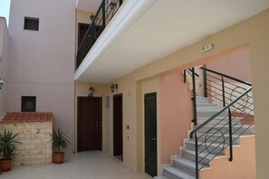 A narrow hallway with a staircase on the right, leading to an upper floor. There are two doors on the left, one of which is open. The walls are painted in a light color, and there are potted plants at the entrance.