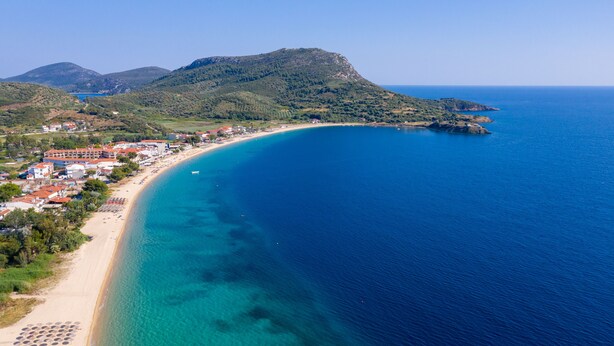Vista aérea de un pueblo costero con una larga playa de arena, aguas azules cristalinas y montañas al fondo.