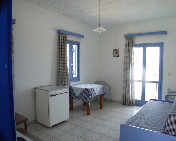A small, simple room with a kitchenette, a table with a checkered tablecloth, and two windows with curtains. - Thalia Apartments (Malevizi)