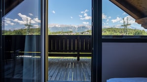 Habitación con ventanas amplias que ofrecen vista a la montaña, una terraza de madera en el exterior y una cama con cubrecama blanca.