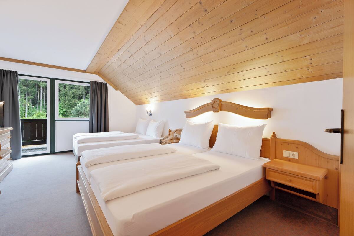 A bedroom with three beds, wooden ceiling beams, a nightstand, and a view of greenery outside.