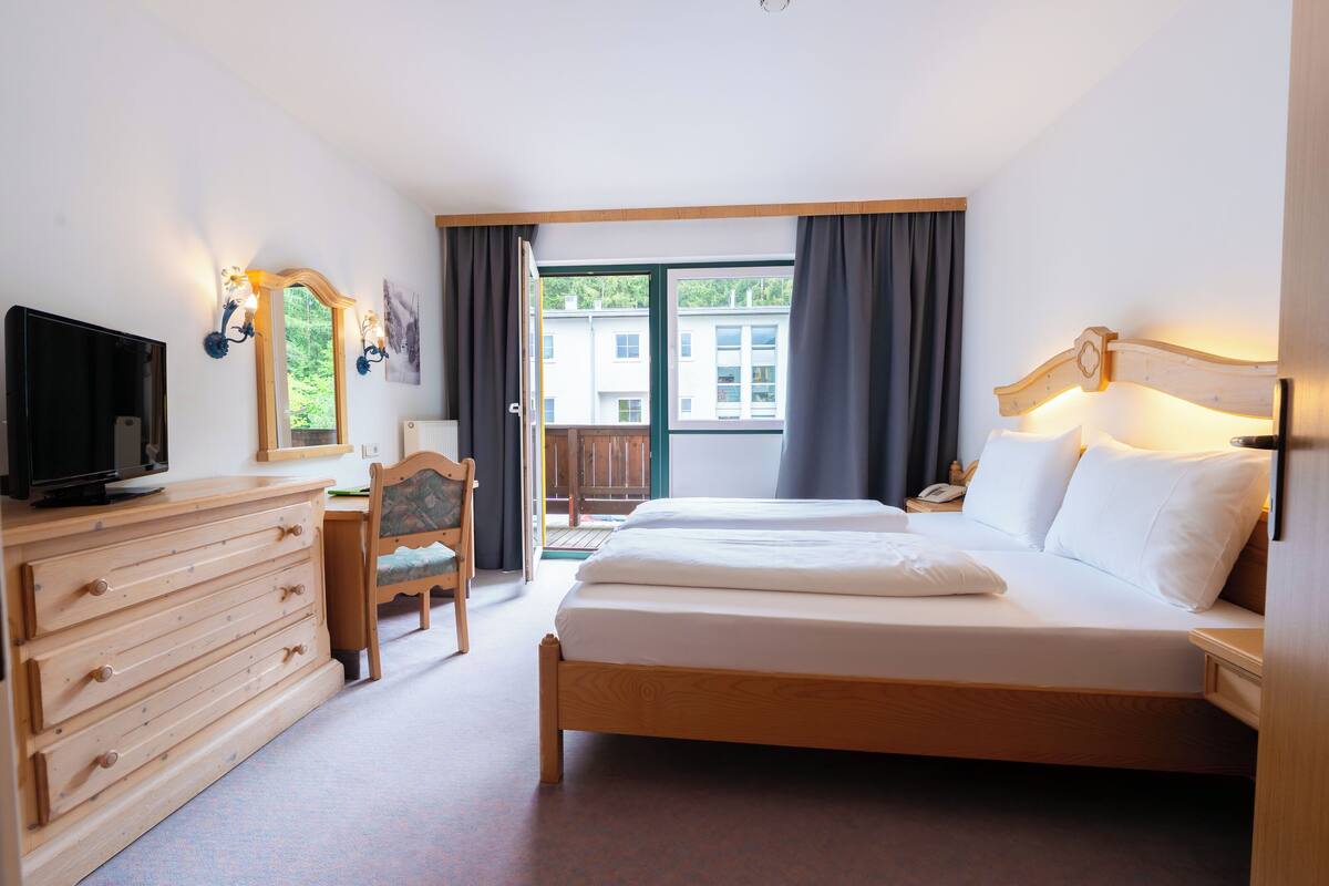 A hotel room with a bed, a desk, a chair, a dresser, a television, and a view of buildings through the window.
