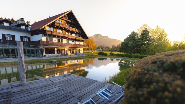 Una terraza de madera con sillones de descanso, un edificio con balcones y un estanque tranquilo que refleja la estructura.