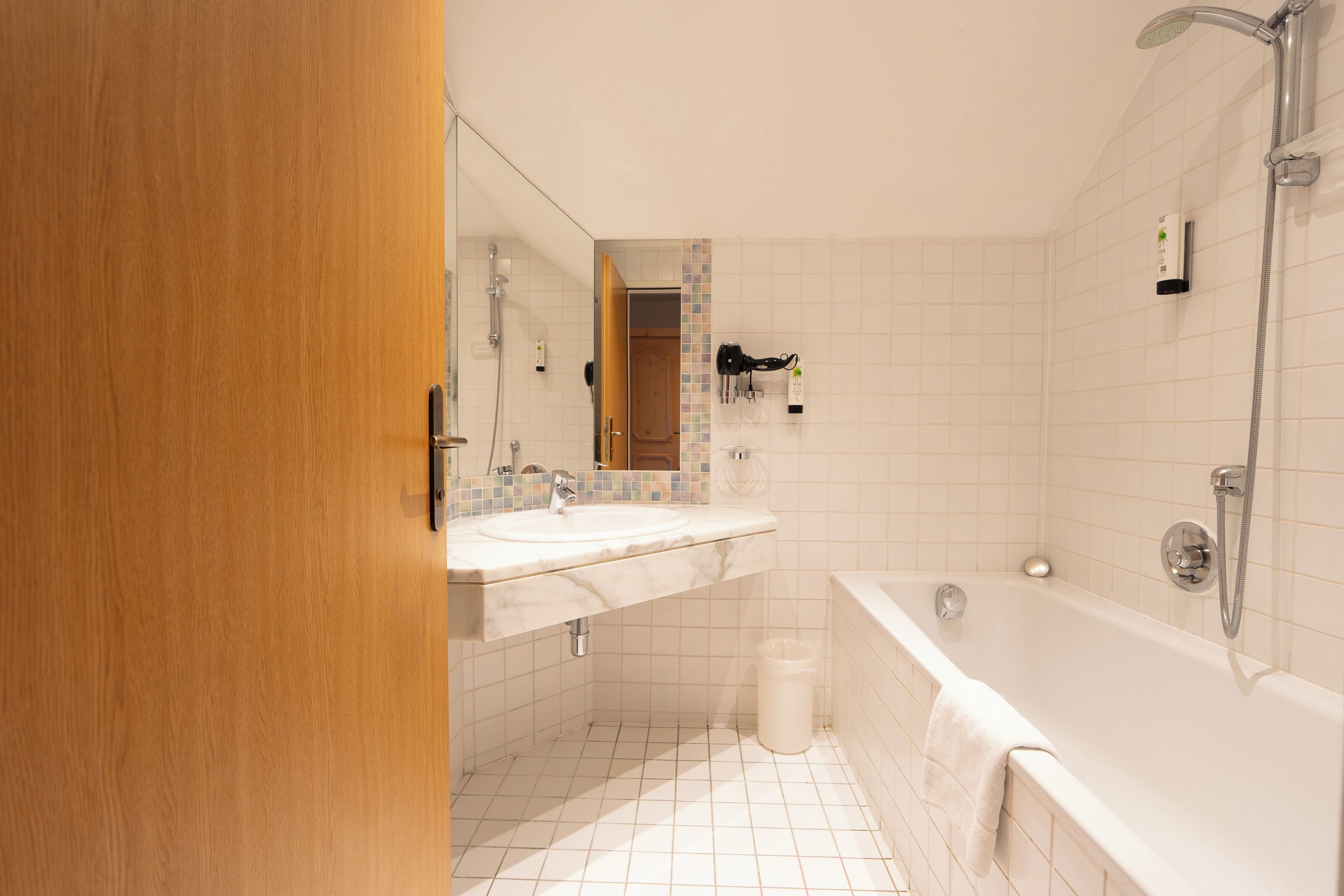 Un baño con puerta de madera, lavamanos blanco, espejo, ducha y cubo de basura blanco.