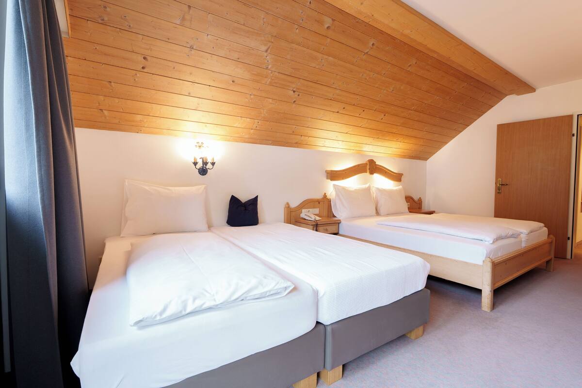 A bedroom with two beds, wooden ceiling beams, a nightstand with a lamp, and a door.