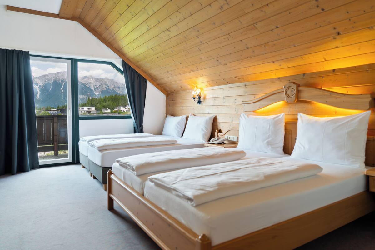A room with three beds, wooden ceiling, large windows, and a view of mountains.
