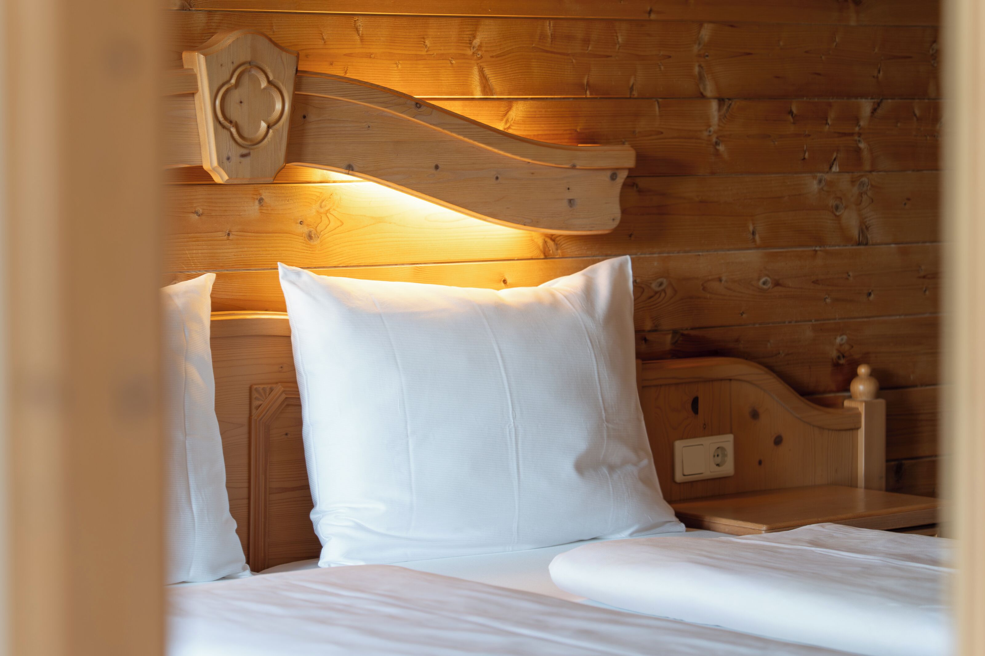 A wooden bed with white bedding, a wooden headboard, and a bedside table with a lamp.