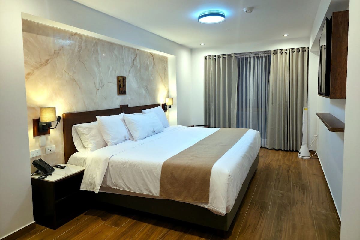 Habitació d'hotel amb un llit gran, tauletes de nit, un telèfon, televisió penjada a la paret i una làmpada al sostre.