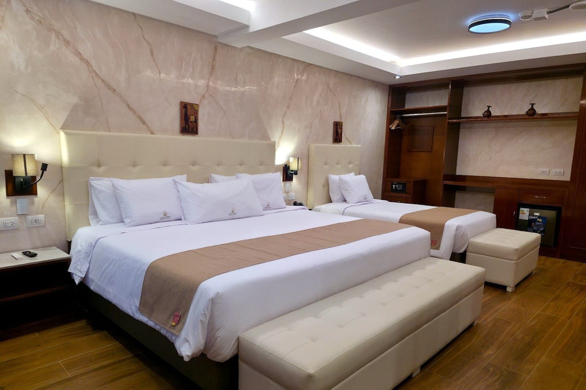 Habitació d'hotel amb un llit gran, una taula de nit, una banca, un armari i un frigorífic petit.