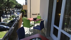 Un balcón con una mesa roja, dos sillas verdes y una ventana con vista a coches estacionados y árboles.