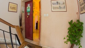Una escalera con barandilla de madera que lleva a una puerta roja con un letrero. Hay cuadros en las paredes y una planta en maceta a la derecha.