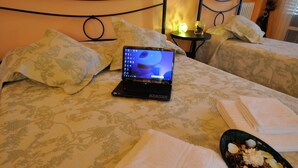 Una laptop sobre la cama, con una colcha estampada, una mesita con lámpara, y toallas dobladas ordenadamente.