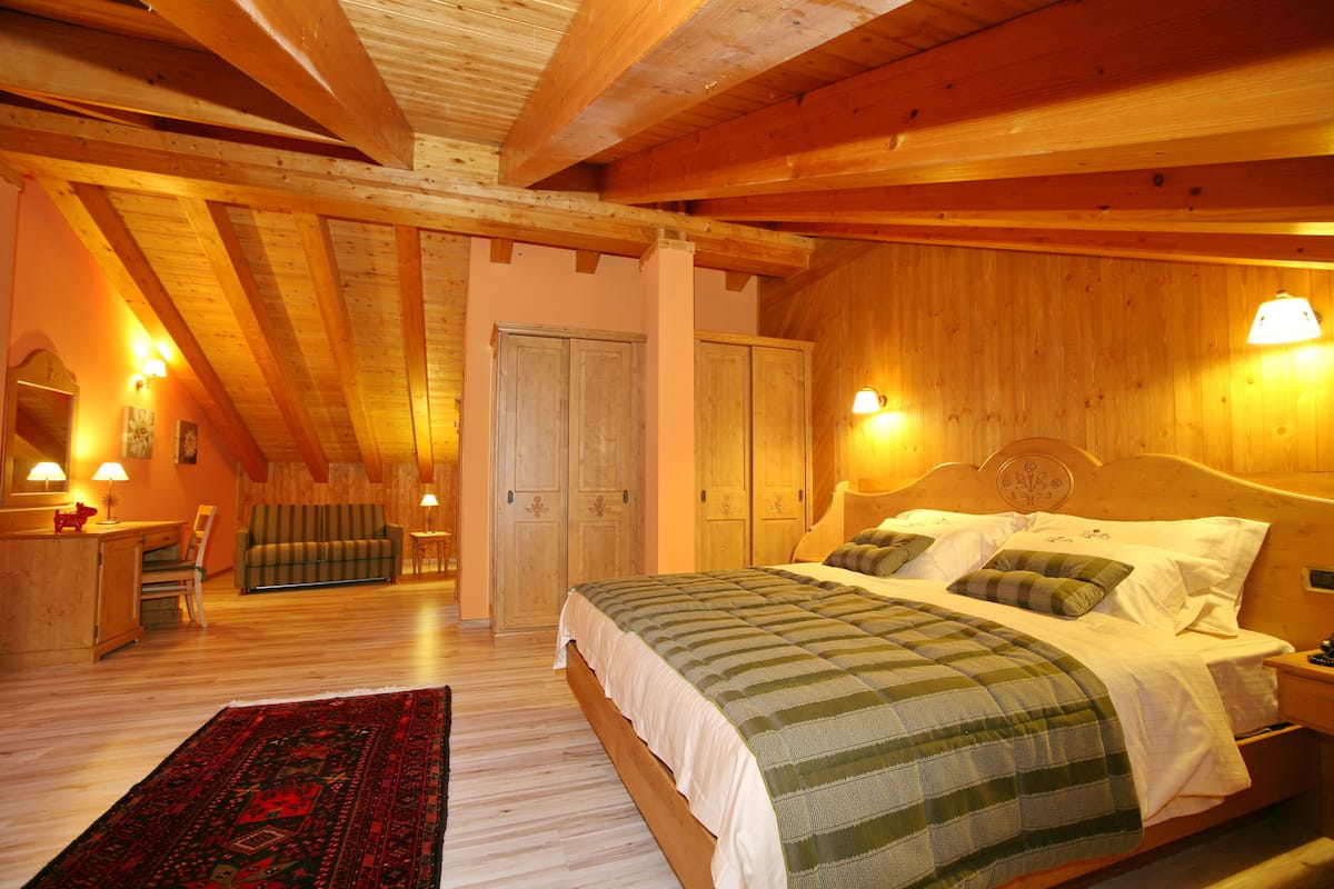 A cozy bedroom with a wooden ceiling, a large bed with green and white bedding, a nightstand, and a red and black patterned rug.