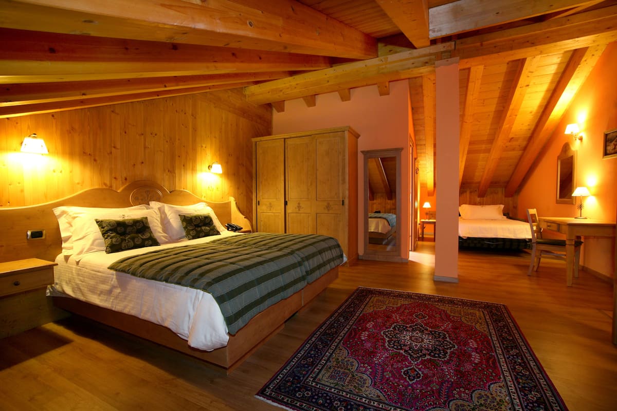 A cozy bedroom with a wooden ceiling, a large bed with a green and white bedspread, a red and blue patterned rug, and a wooden wardrobe.