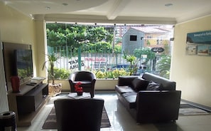 A living room with a sofa, armchairs, a TV, and a view of a city street with greenery and a signboard.