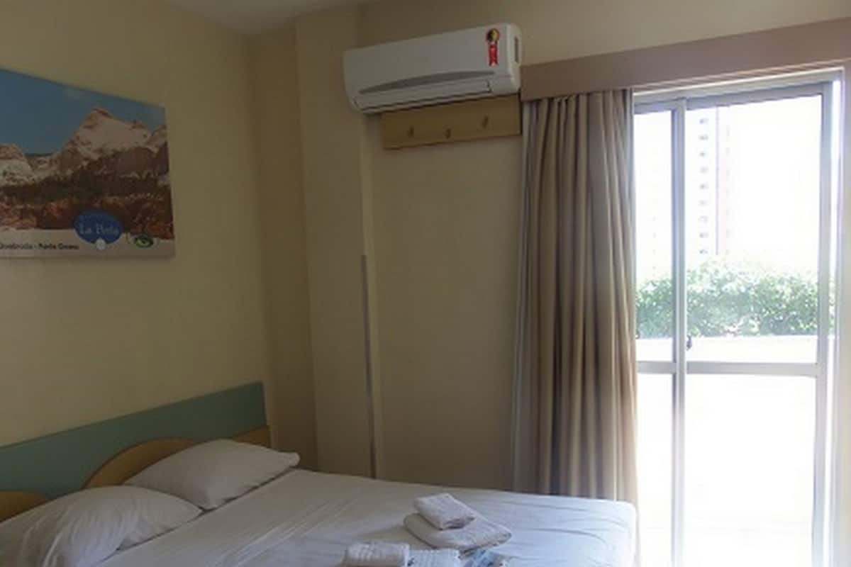 Una habitación de hotel con cama, unidad de aire acondicionado, ventana con cortinas y un cuadro en la pared.