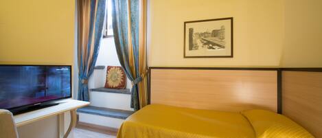 Habitación de hotel con cama, televisor en un escritorio, ventana con cortinas y un cuadro en la pared.