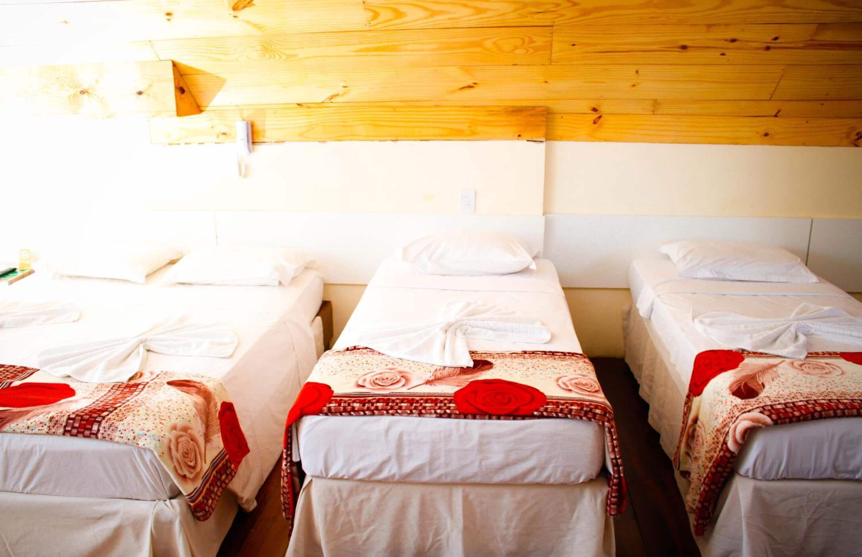 A room with three single beds, each with a white bedspread and red rose decorations. The room has wooden walls and a wooden ceiling.