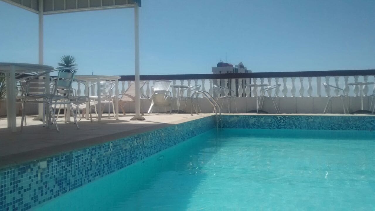 A rooftop pool area with clear blue water, surrounded by white lounge chairs and tables.