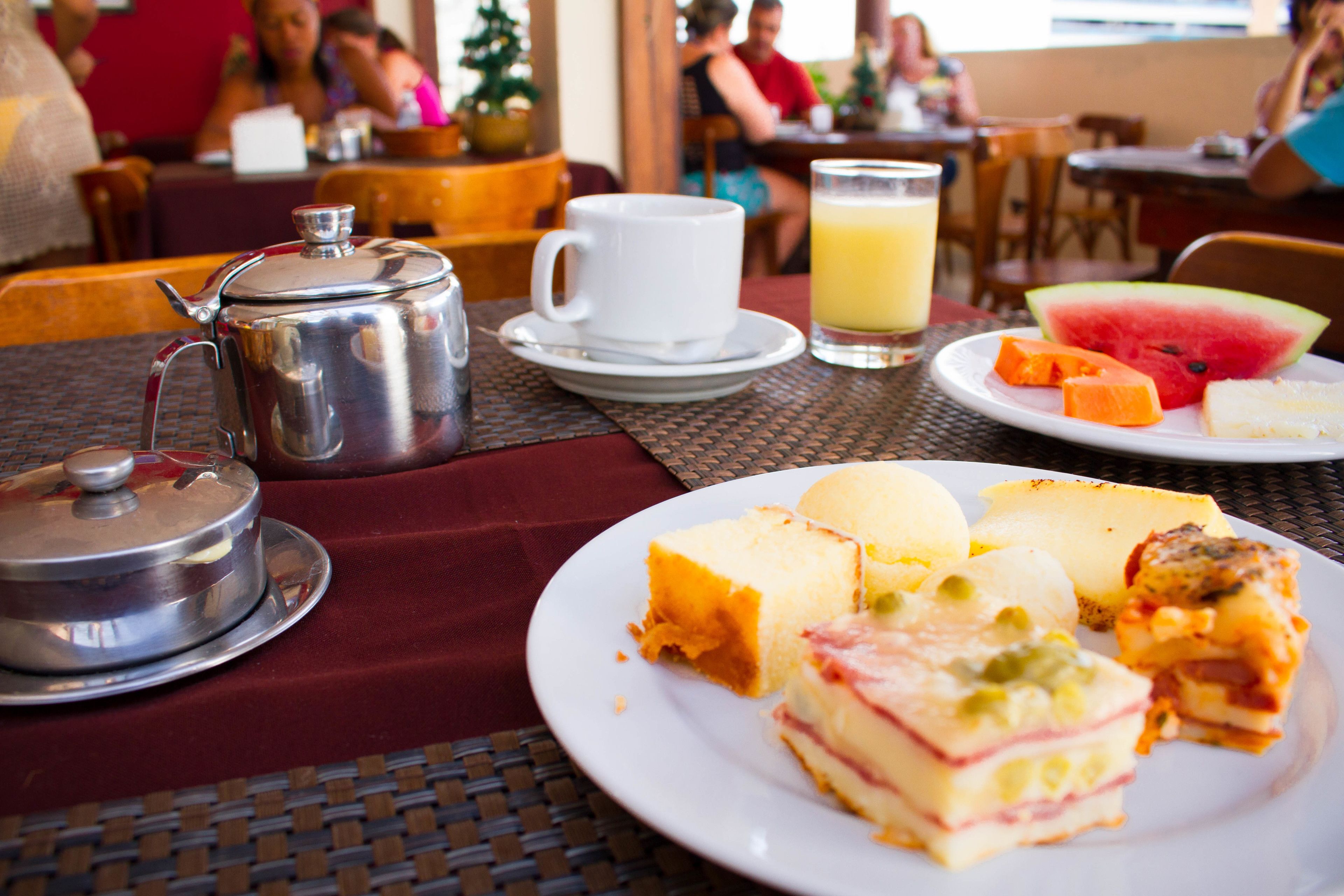 A breakfast setting with a metal coffee pot, white cup, glass of orange juice, and plates of food including a slice of watermelon and cheese.