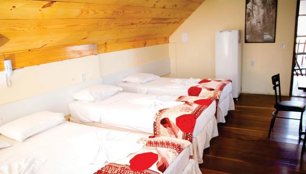 A hotel room with four beds arranged in a row, each with white bedding and red accents. There is a wooden ceiling and a white wall with a painting.