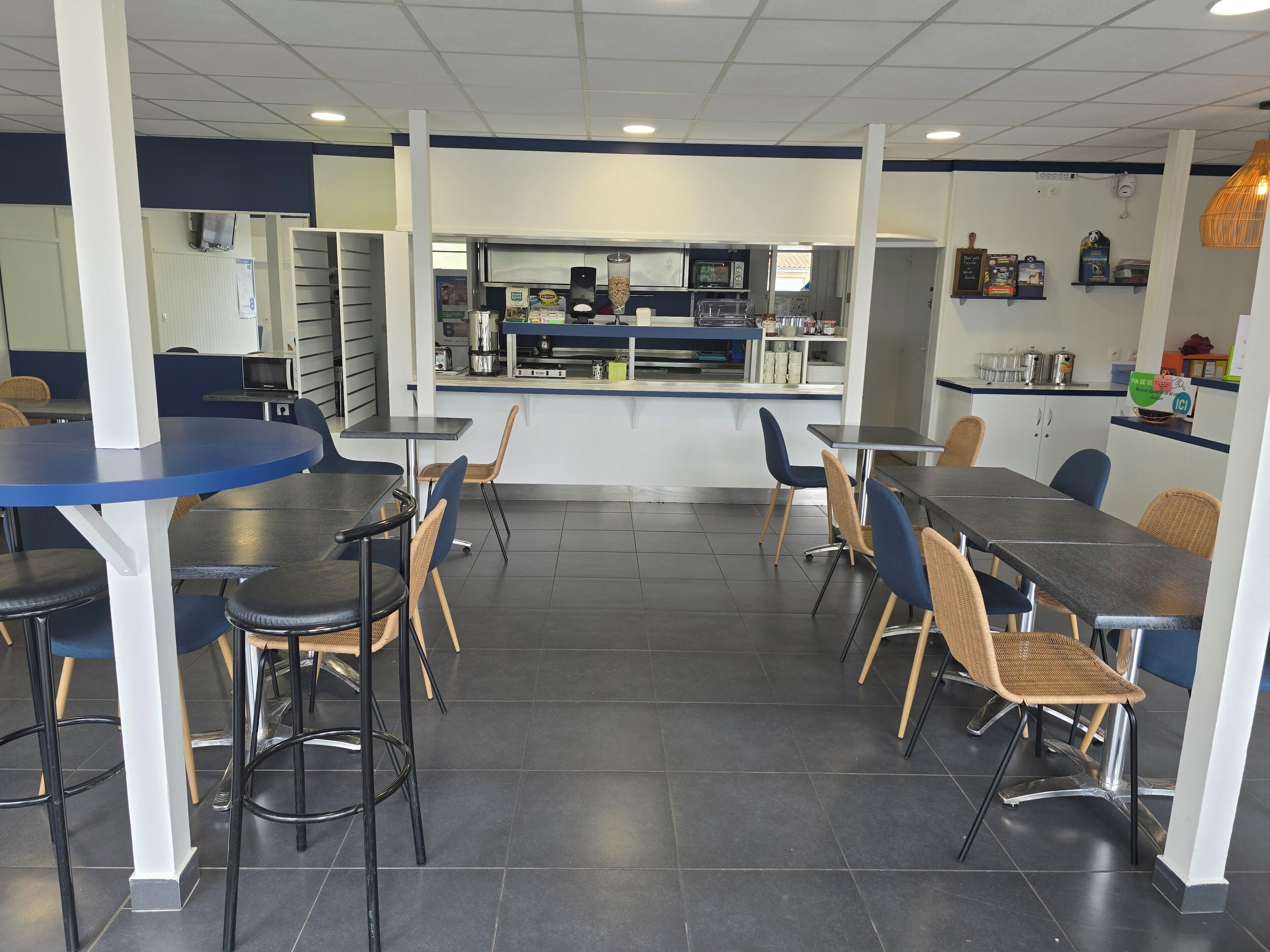 A restaurant with multiple tables and chairs, a counter with a cash register, and kitchen equipment in the background.